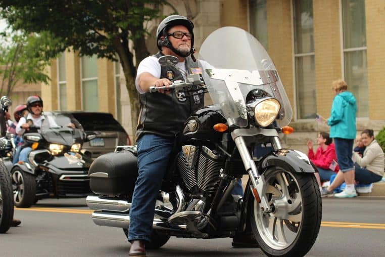 Motard barbu sur une grosse moto lors d’un défilé en ville, roulant calmement et concentré, rappelant les visites fidèles au cimetière.