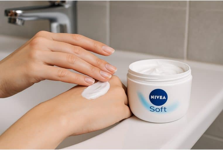Gros plan sur une main féminine appliquant une crème hydratante blanche issue d’un pot rond posé près d’un lavabo, évoquant l’usage d’une crème type Nivea Soft au quotidien.