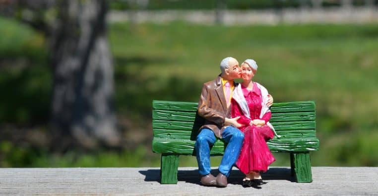 Couple de grands-parents assis enlacés sur un banc dans un parc ensoleillé, illustrant la retraite à deux après une longue vie de travail