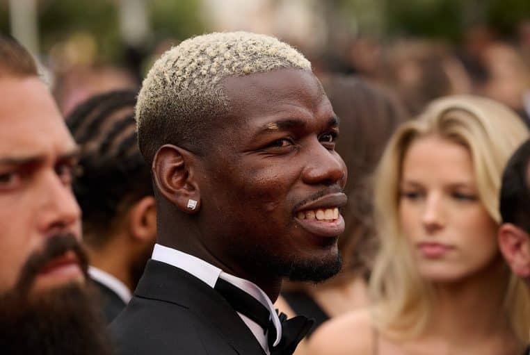 Paul Pogba en costume noir au Festival de Cannes, portrait serré de profil, sourire, foule floue derrière.