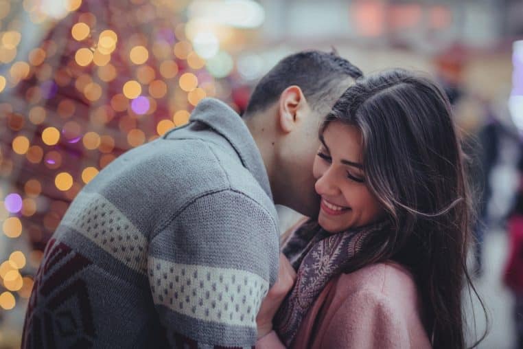 Jeune couple souriant serré l’un contre l’autre devant des lumières de Noël floues, suggérant une rencontre chaleureuse en fin d’année.