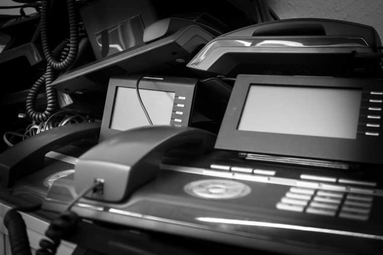 Pile de téléphones de bureau noirs empilés en noir et blanc, symbolisant l’accumulation de vieux appareils électroniques devenus déchets