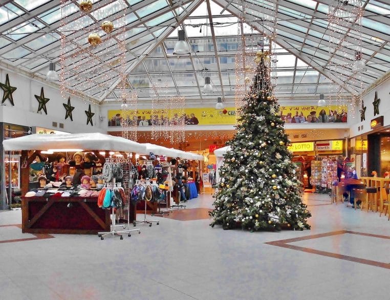 Intérieur d’un centre commercial décoré pour Noël, grand sapin au centre et stands éphémères autour, représentant la concurrence des galeries marchandes.