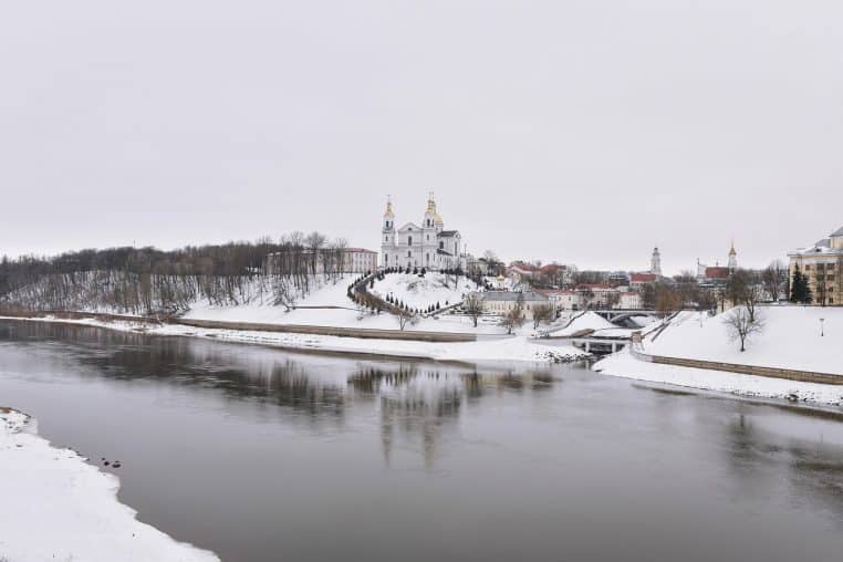 Vue d’une rivière gelée longeant une petite église entourée de neige, sous un ciel d’hiver clair et lumineux dans une ville européenne.