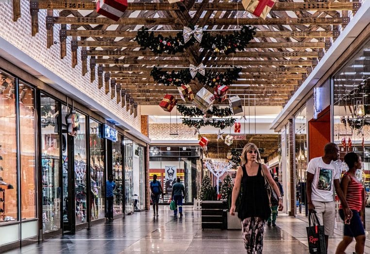 Galerie marchande décorée pour Noël avec cadeaux suspendus au plafond et clients chargés de sacs circulant entre les boutiques.