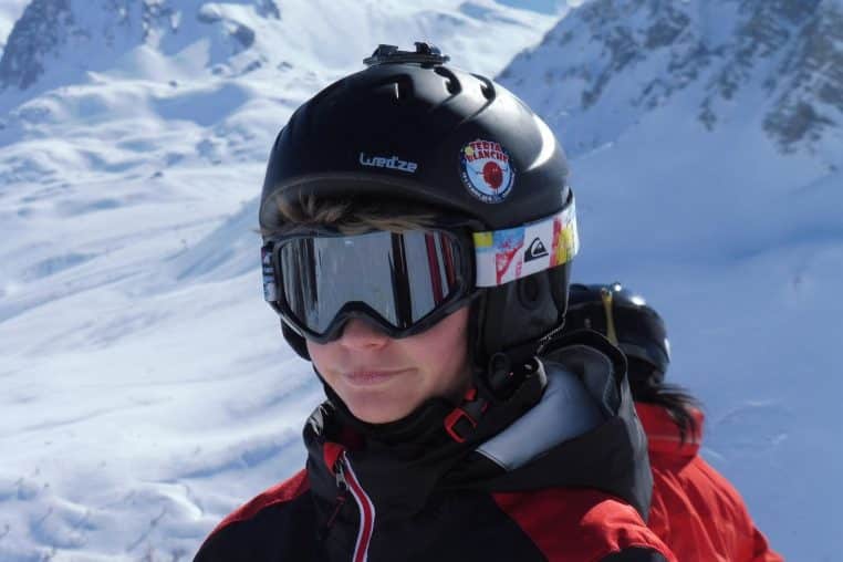 Jeune skieur en veste rouge dévalant une piste enneigée, un ami assis derrière lui rit encore après une chute.