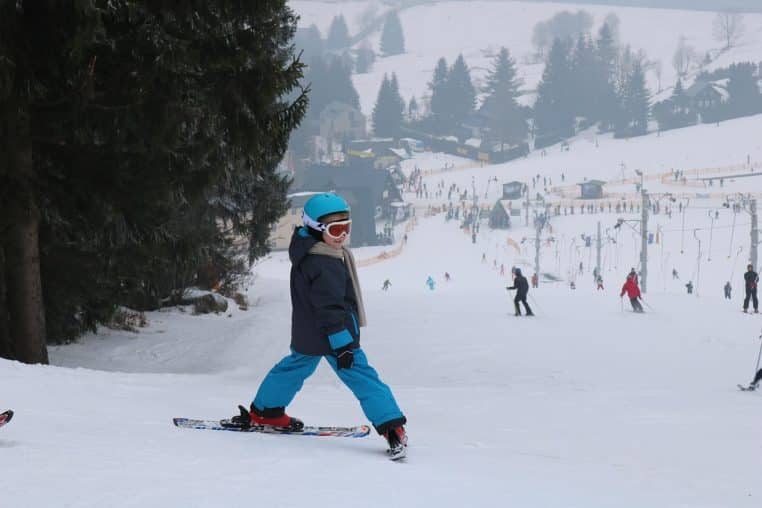Enfant en combinaison de ski colorée debout sur une piste enneigée, casque sur la tête et bâtons à la main devant les reliefs de la station