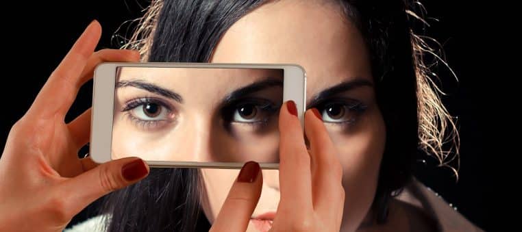 Gros plan sur un smartphone tenu à bout de bras, affichant les yeux d’une femme sur l’écran, suggérant le regard insistant derrière chaque appel.