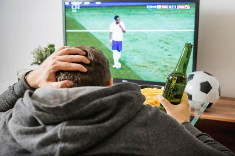 Homme assis dans son salon avec une bière à la main, regardant un match de football sur un grand téléviseur, symbole du piratage des compétitions sportives.
