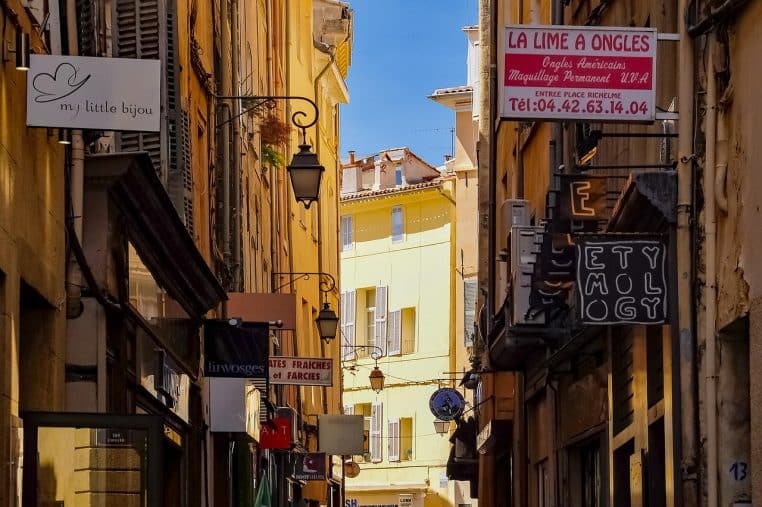 Ruelle commerçante étroite d’une ville du Sud, bordée de petites enseignes et d’enseignes suspendues, baignée de lumière en plein après-midi.