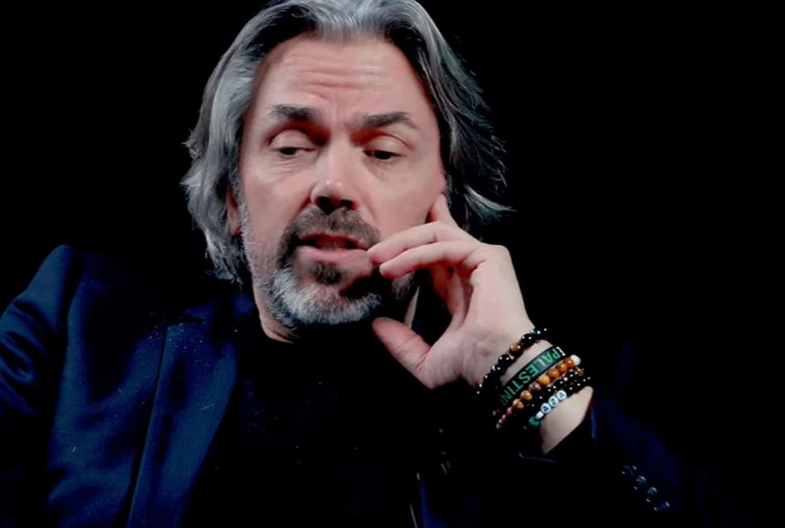 Un homme aux cheveux grisonnants et barbe courte, filmé en gros plan sur fond noir pendant une interview filmée.