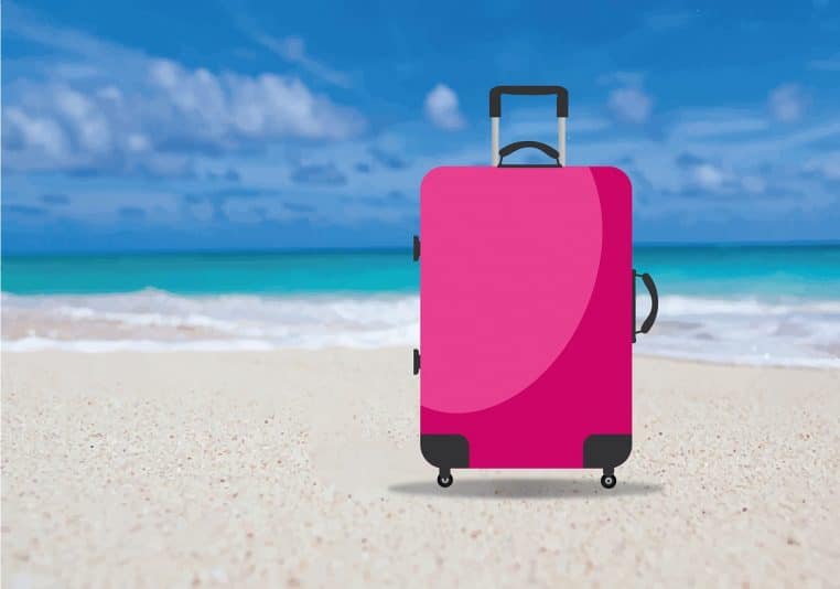 Grande valise rose posée sur une plage de sable devant la mer turquoise et le ciel bleu, évoquant un départ en congés payés.
