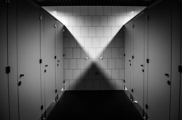 Rangée de cabines de toilettes dans un couloir carrelé, éclairage contrasté qui forme un triangle de lumière au fond de la pièce.