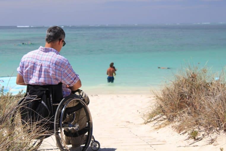 Personne en fauteuil roulant contemplant la mer depuis un chemin de sable, face à une plage accessible et des baigneurs au loin.