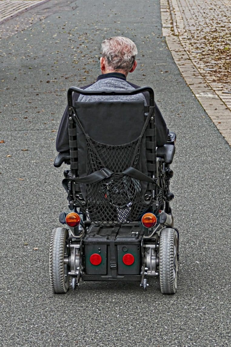 Homme vu de dos dans un fauteuil roulant électrique circulant seul sur une allée goudronnée bordée de pavés et d’arbres.
