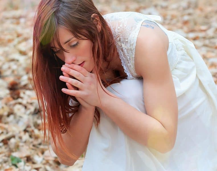 Jeune femme aux cheveux roux, mains jointes et yeux clos, agenouillée dehors, comme recueillie devant une tombe ou un souvenir.