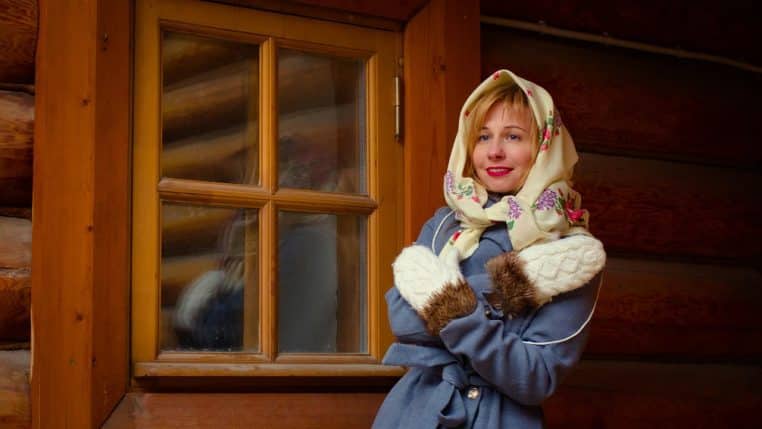 Jeune femme emmitouflée dans un manteau d’hiver devant une maison en bois, fenêtres vitrées visibles sous la neige