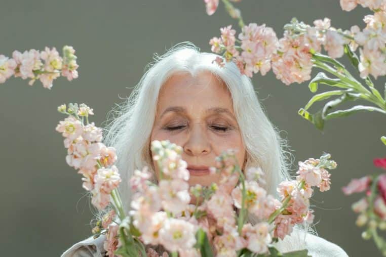 A peaceful elderly woman with white hair among pink blossoms, eyes closed, enjoying tranquility.