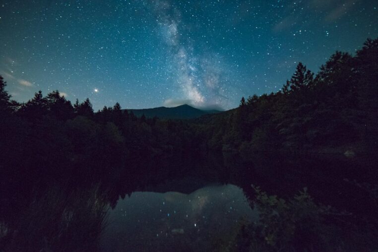 A picturesque night sky with Milky Way over a serene mountain and forest landscape.