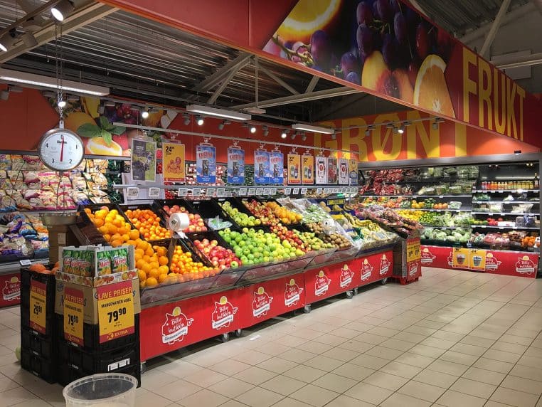 Étal de fruits et légumes dans un supermarché, bacs colorés et panneaux promotionnels au-dessus, vue panoramique.