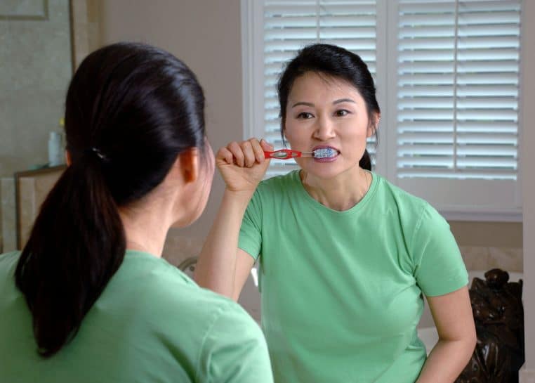 Femme se brossant les dents face à un miroir, illustration concrète des gestes d’hygiène bucco-dentaire.