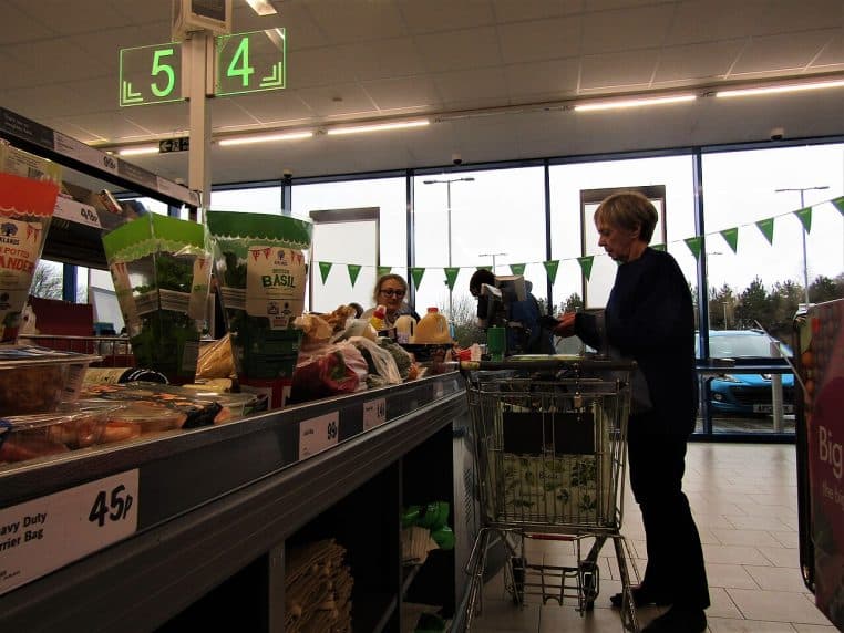 Caisse de supermarché avec tapis roulant chargé, chariot et client au paiement, lumière naturelle en vitrine.