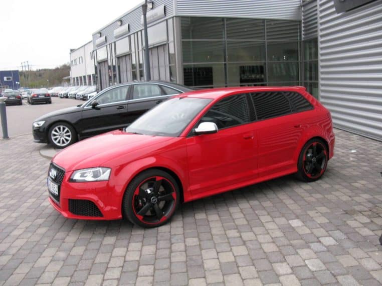 Audi RS3 rouge garée devant un bâtiment, vue trois-quarts avant, jantes sport et carrosserie brillante.
