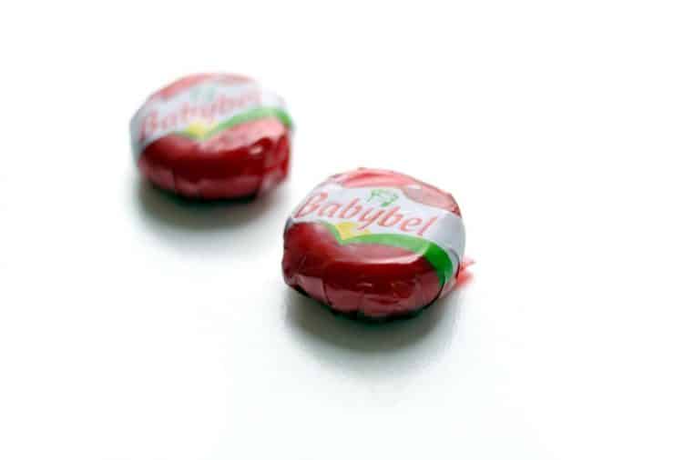 Deux Mini Babybel emballés posés sur une table, cire rouge intacte et filet visible en arrière-plan.