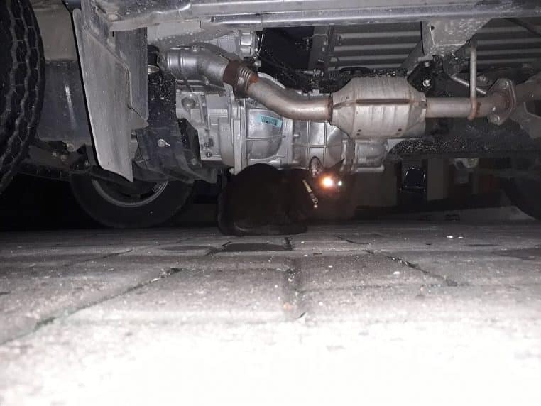 Chat noir caché sous une voiture, sol pavé, flash lumineux au premier plan et dessous de châssis visible.