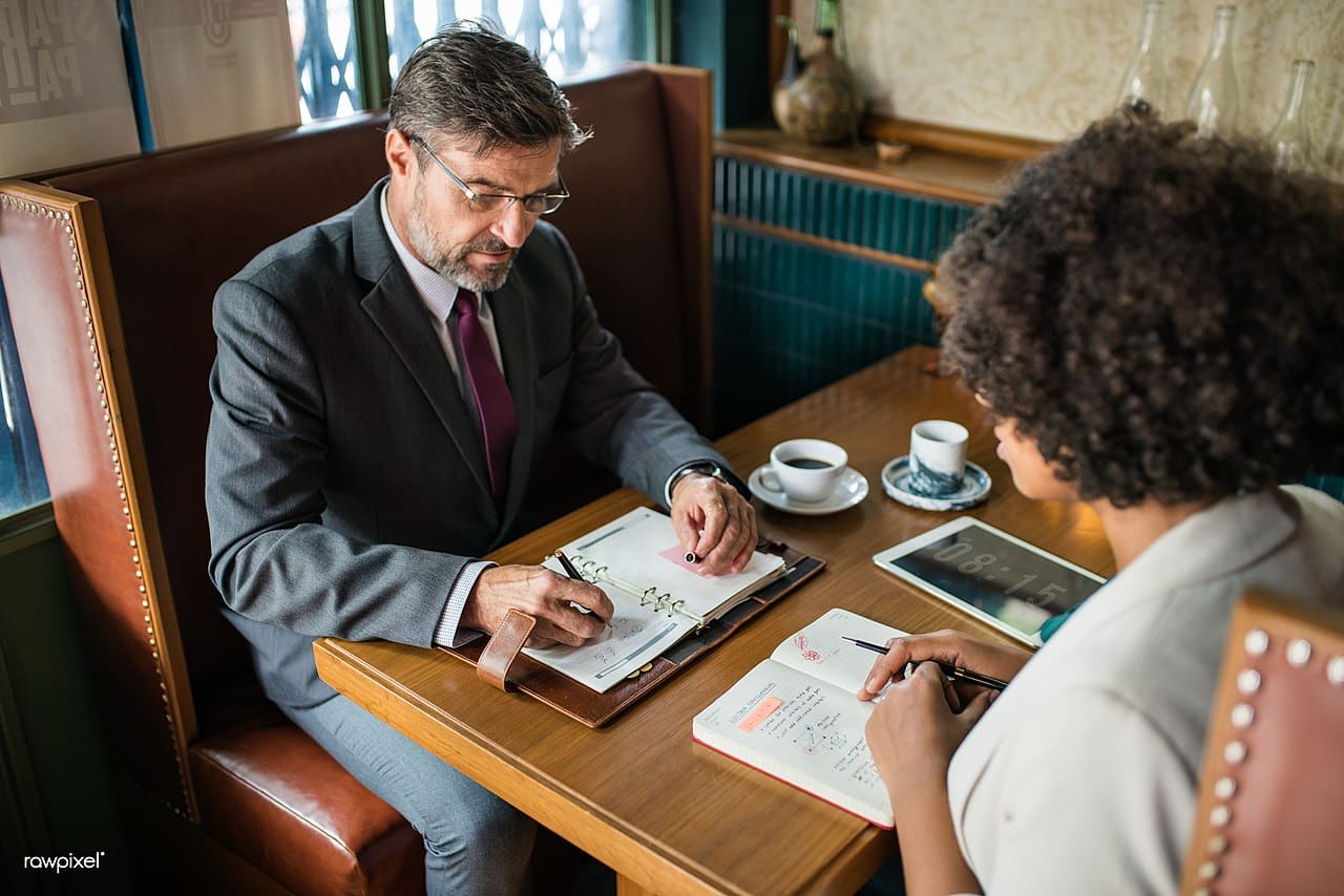 Deux collègues discutent dans un café, carnet de notes ouvert sur la table.