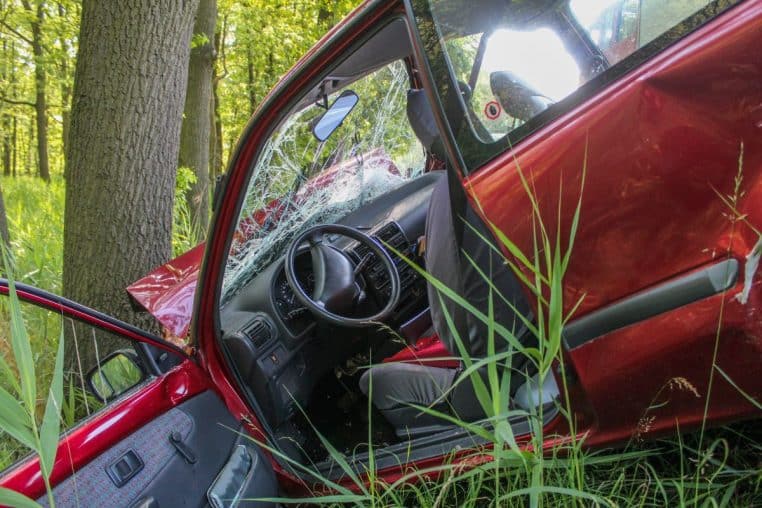 Intérieur d’une voiture accidentée près d’un arbre, pare-brise fissuré et portière ouverte en plein jour.