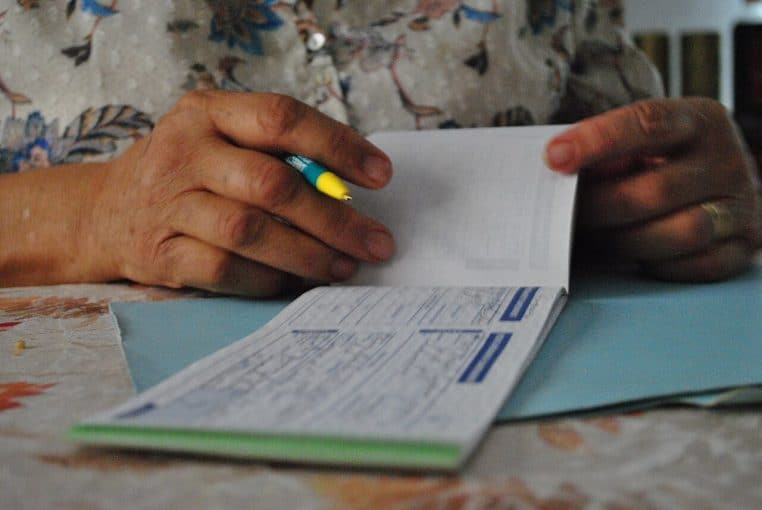 Une personne âgée écrit un chèque sur une table, geste de paiement et documents posés à proximité