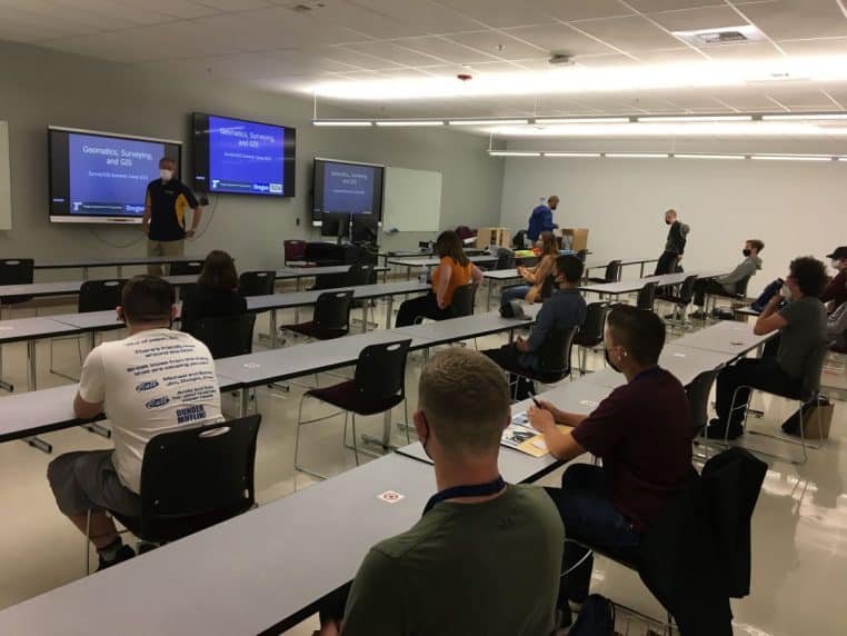 Salle de cours avec participants assis à des tables, regard tourné vers l’intervenant et écrans de présentation.