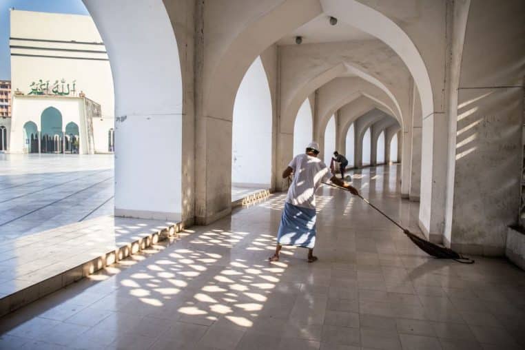 Personne en train de balayer et nettoyer un sol en intérieur, grande allée lumineuse et carrelage clair.