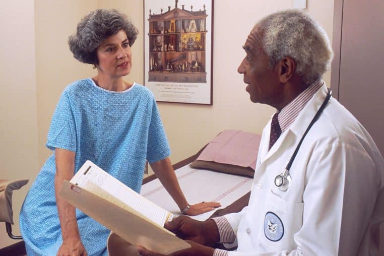 Médecin en blouse discutant avec une patiente assise sur une table d’examen, dossier médical à la main, scène de consultation.