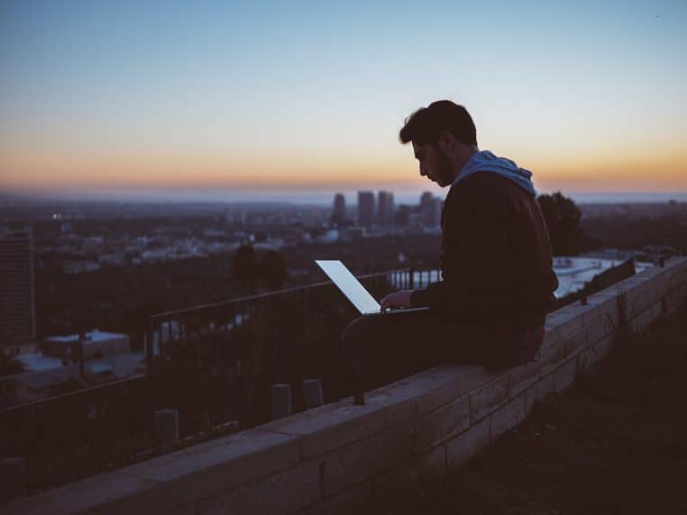 Jeune homme travaillant sur un ordinateur portable face à une baie vitrée, lumière de coucher de soleil en ville.