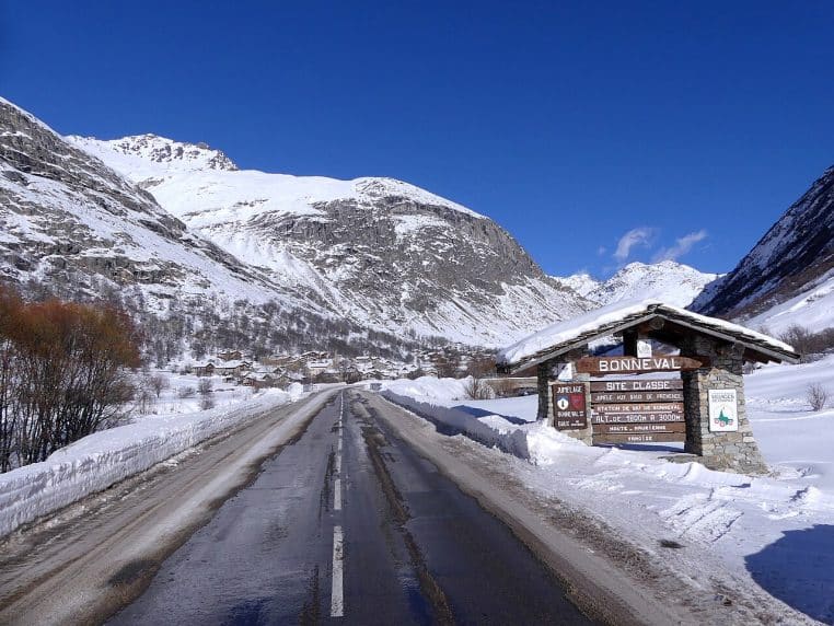 Entrée de Bonneval-sur-Arc sous la neige, route dégagée au centre et montagnes blanches sous ciel bleu.