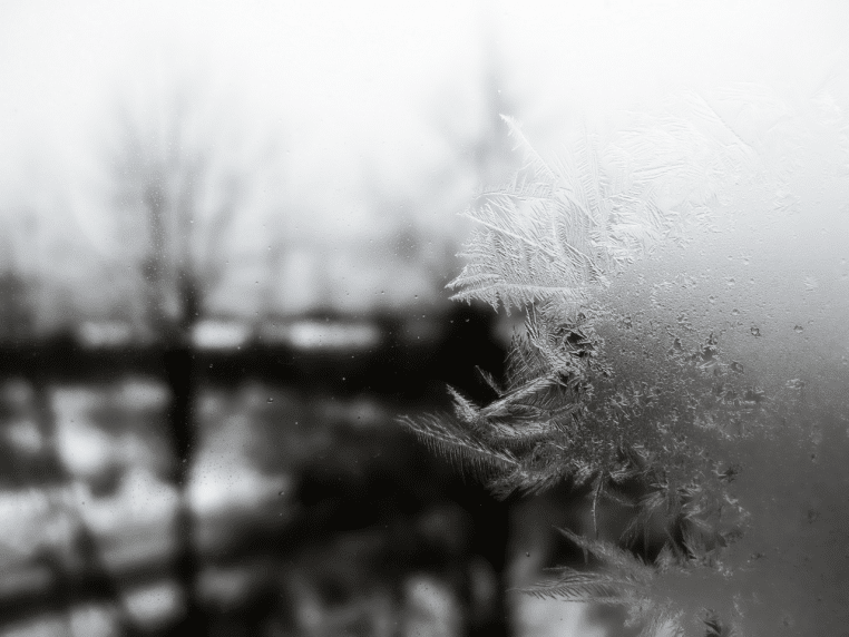 Fenêtre couverte de givre, cristaux de glace au premier plan et jardin enneigé flouté en arrière-plan.