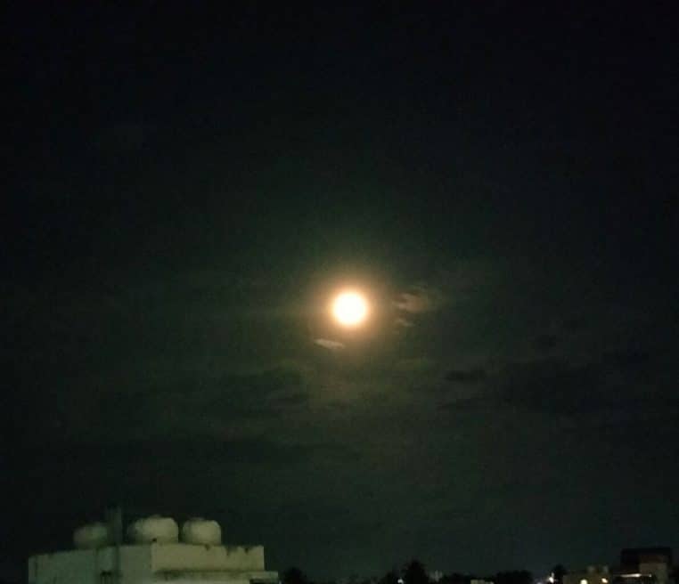 Pleine lune ronde dans un ciel nocturne légèrement voilé, halo lumineux au-dessus des toits d’une ville.