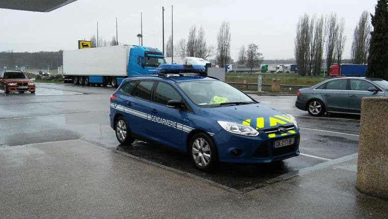 Voiture de gendarmerie bleue stationnée sur une aire routière par temps de pluie, gyrophares visibles, camions en arrière-plan.