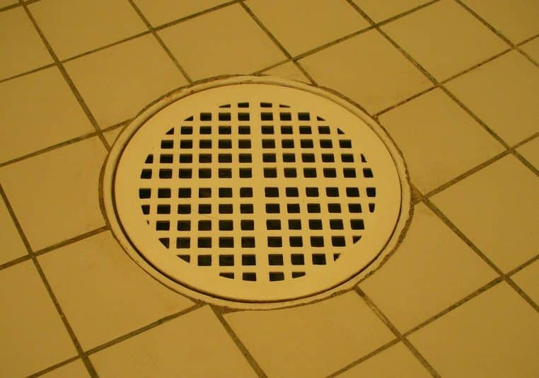 Gros plan d’une bonde de douche au sol, grille métallique et carrelage autour, prête à être nettoyée.