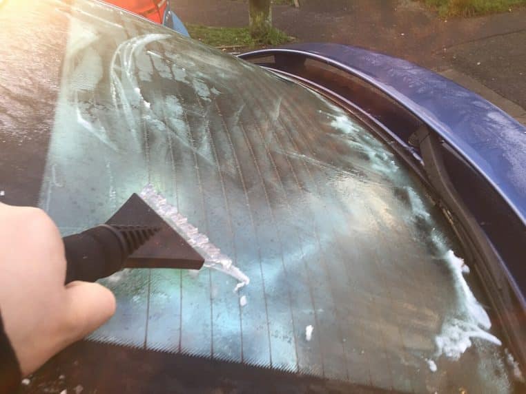 Main utilisant un grattoir à glace sur un pare-brise gelé, avec des traces de dégivrage visibles sur la vitre.