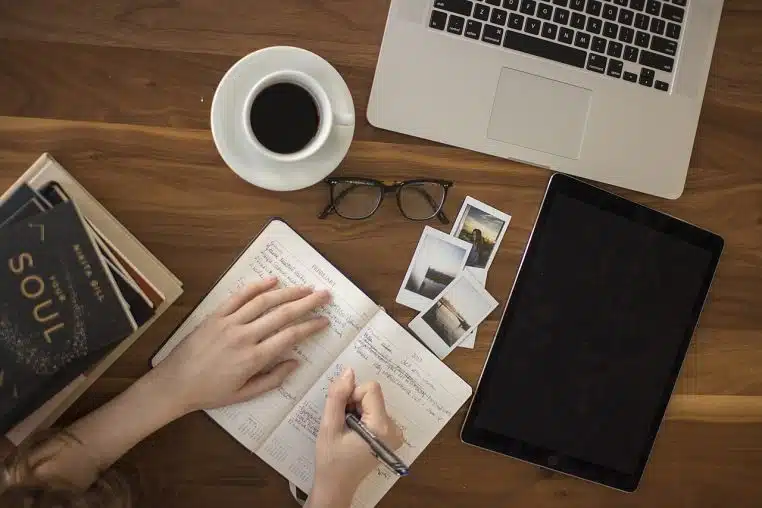 Mains écrivant dans un carnet sur une table en bois avec café, lunettes et tablette, moment de pause.