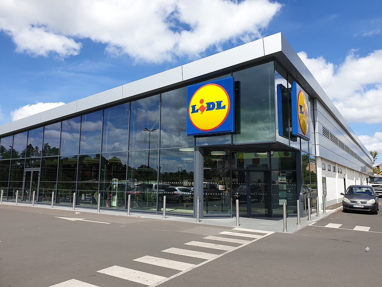 Magasin Lidl moderne avec grande façade vitrée, logo en angle, passage piéton au premier plan, ciel bleu.