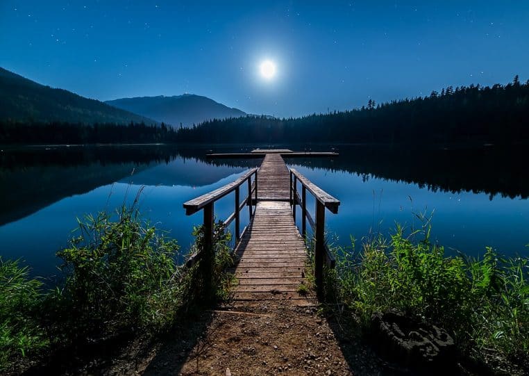 Pleine lune brillante au-dessus d’un lac calme, ponton en bois au premier plan et montagnes sombres au loin.