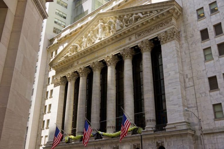 Façade monumentale de la New York Stock Exchange, colonnes et fronton sculpté, drapeaux américains au premier plan.