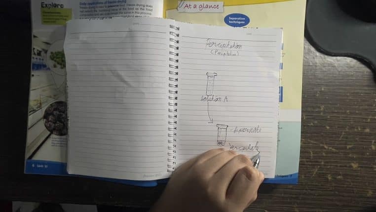 Main écrivant dans un carnet ouvert sur un bureau, pages lignées et croquis, ambiance studieuse et calme.