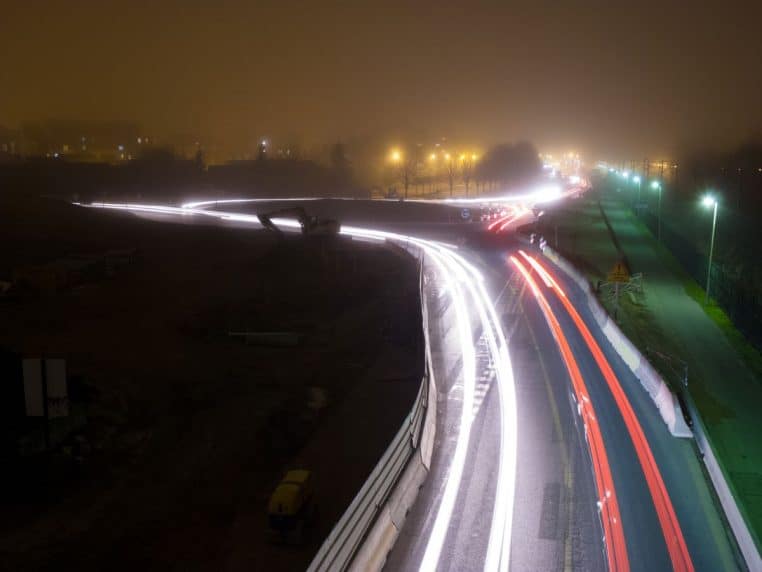 Rond-point vu de haut de nuit, traînées lumineuses de phares et brouillard, circulation sur plusieurs voies.