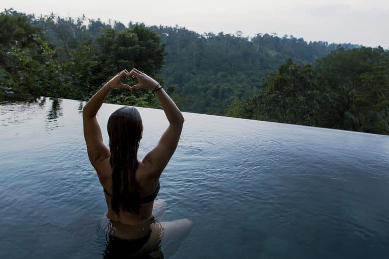 Femme méditant dans une piscine à débordement face à une forêt, bras levés en cœur, ambiance calme.