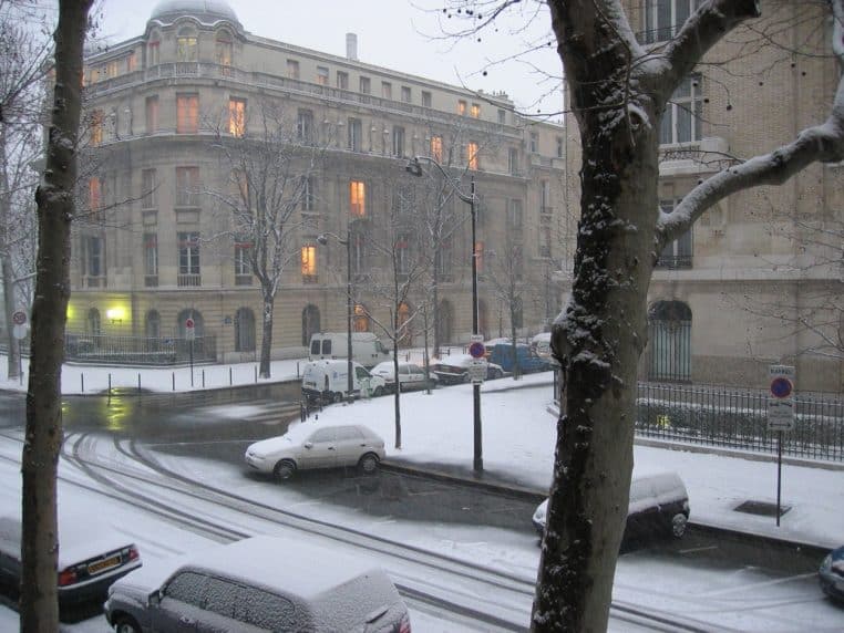Scène de rue à Paris sous la neige, avec chaussée blanchie et passants emmitouflés.